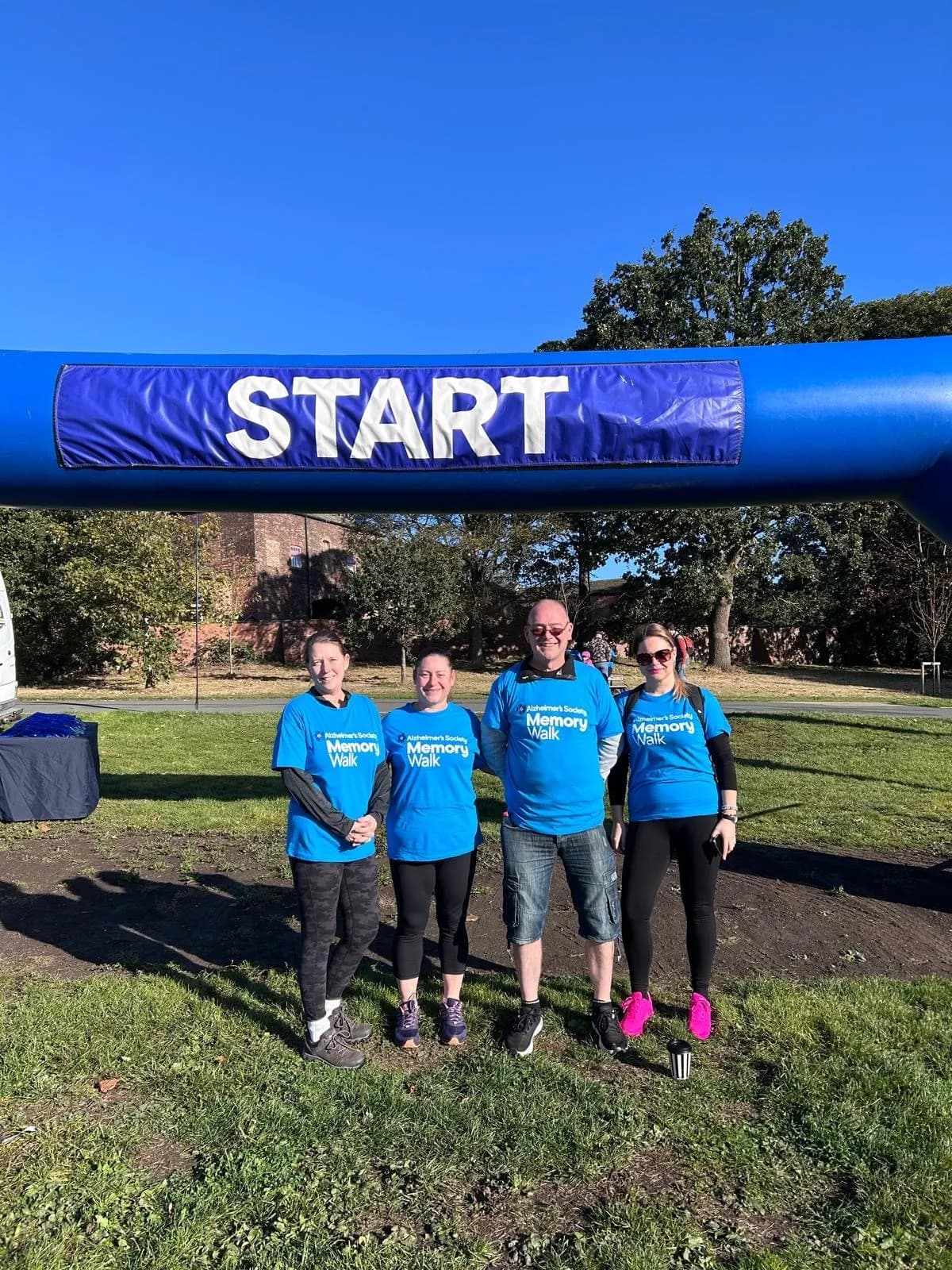 Four people in blue t-shirts that say "Memory Walk" stand under an inflatable arch that says "START" on a sunny day.